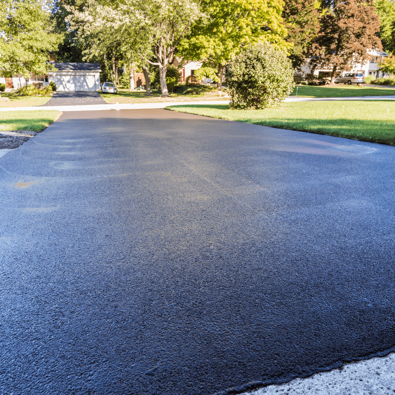 Untitled-design-21 Freshly paved black asphalt driveway in a suburban neighborhood with green lawns and trees.