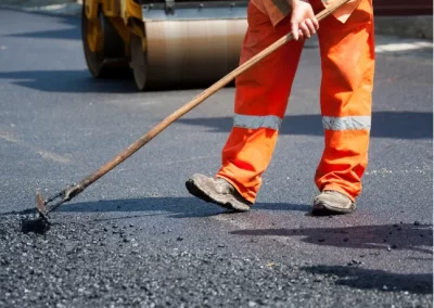 Worker in orange safety pants leveling fresh asphalt with a rake near a road roller, showcasing the precision of Jones Asphalt VA Expert Asphalt Services.