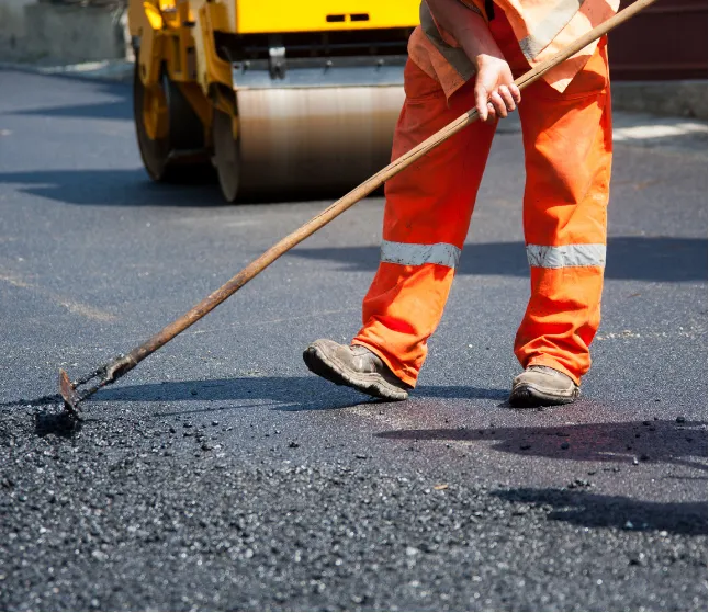 asphalt-maintenance-1 Construction worker in orange gear spreading asphalt on a road with a rake; roller machine in background.
