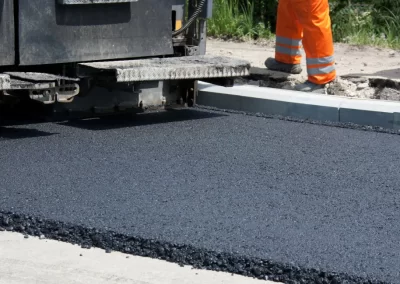 Fresh asphalt being laid by a paving machine as a worker in orange pants stands nearby, showcasing Jones Asphalt VA Expert Asphalt Services at work.