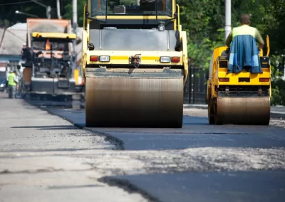 Two road rollers flatten fresh asphalt on a city street while Jones Asphalt VA Expert Asphalt Services workers operate machinery in the background.