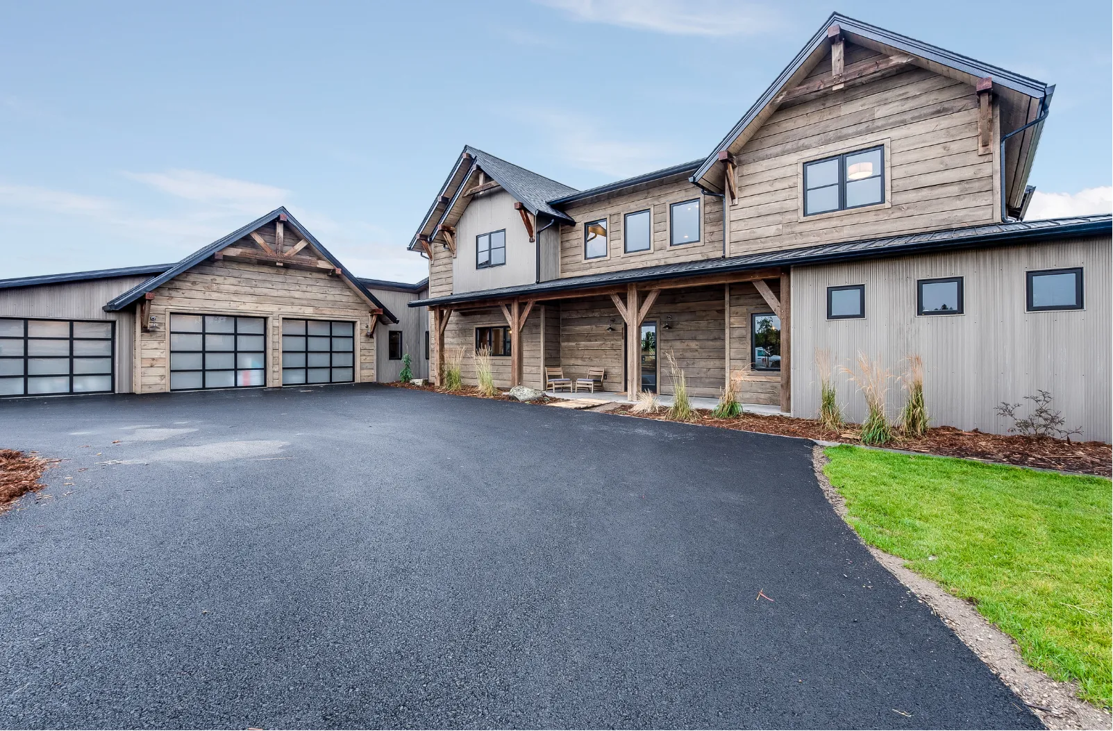driveway-h Modern rustic house with wood siding, large windows, attached garage, and paved driveway on a clear day.