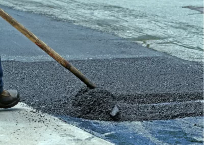 A worker from Jones Asphalt VA spreads fresh asphalt on a road with a shovel; an orange traffic cone is visible in the background, showcasing expert asphalt services.