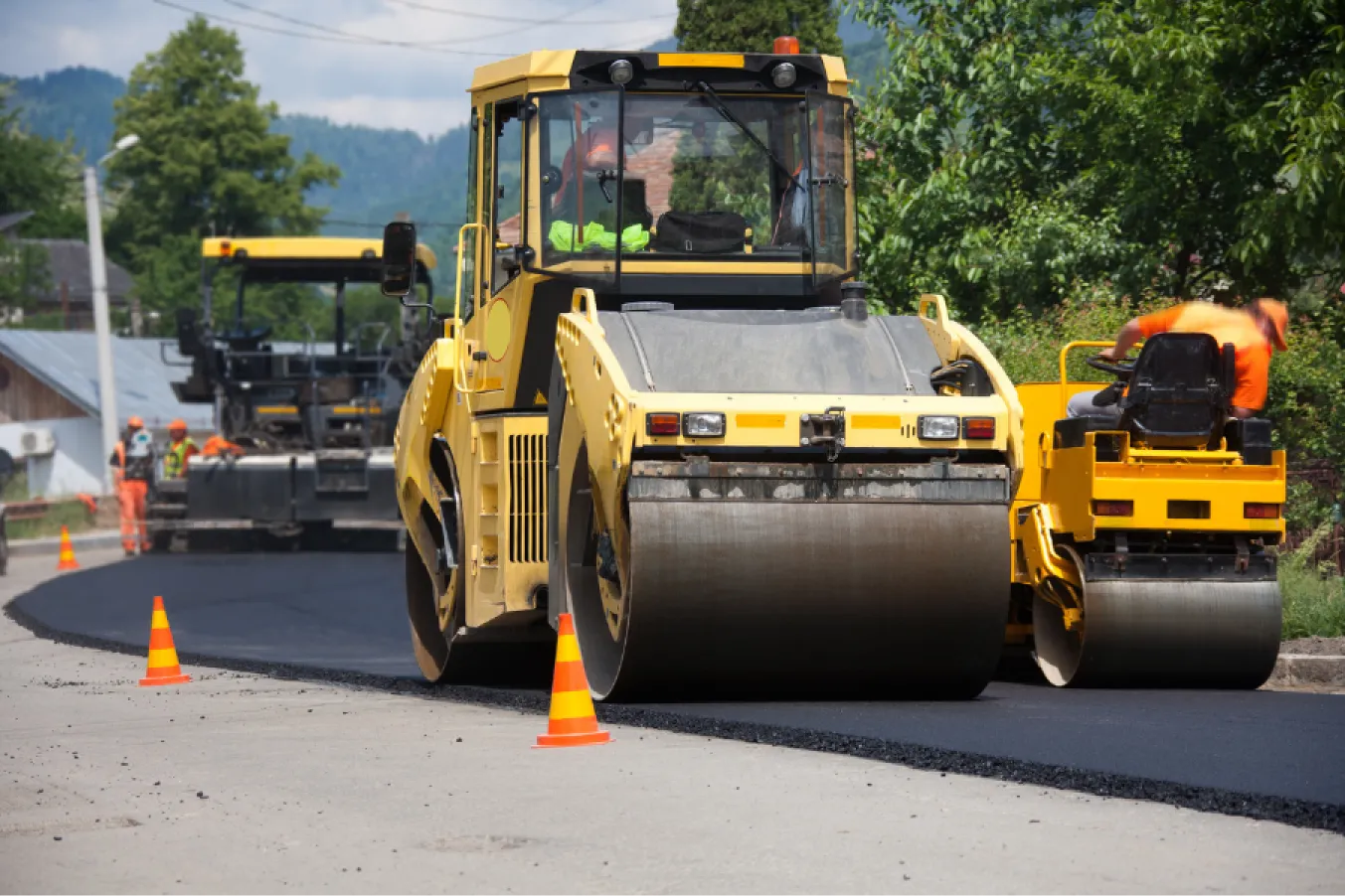 about-1 Road construction with yellow steamrollers flattening fresh asphalt, outlined by orange cones on a sunny day.