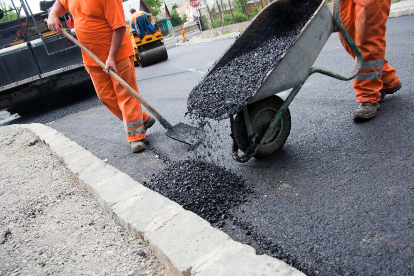 about-2 Workers in orange uniforms pour and spread asphalt from a wheelbarrow onto a road under construction.