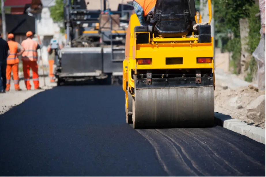 A steamroller flattens fresh asphalt while workers in orange vests work on a new road.