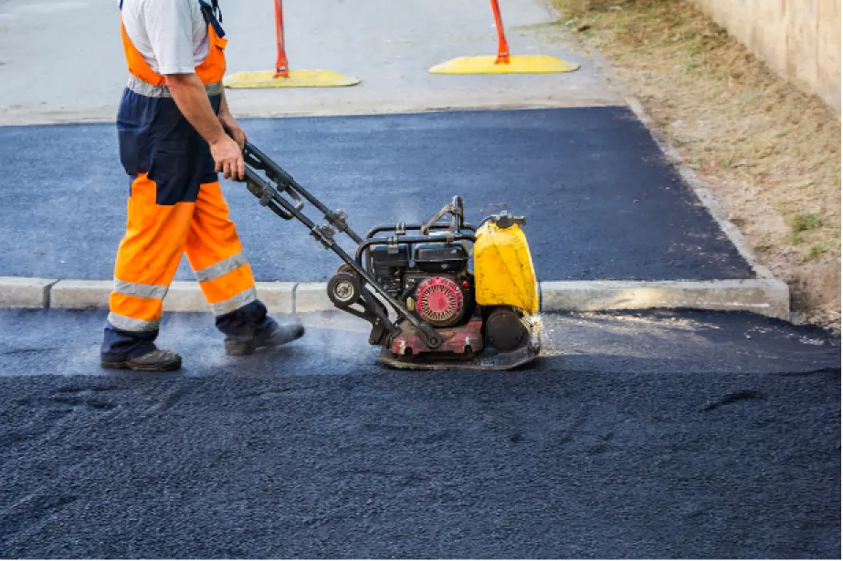 A worker uses a compactor machine to flatten freshly laid asphalt on a road.