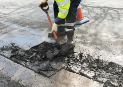 Jones Asphalt VA Expert Asphalt Services worker in high-visibility gear uses a shovel to repair a damaged section of road near a traffic cone.