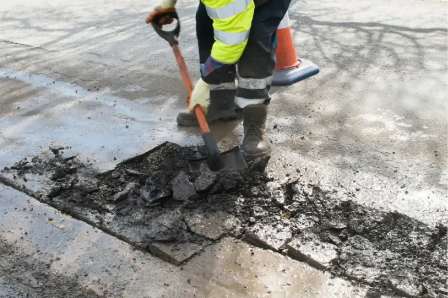 Worker in safety gear uses a shovel to repair a damaged section of asphalt road near a traffic cone.