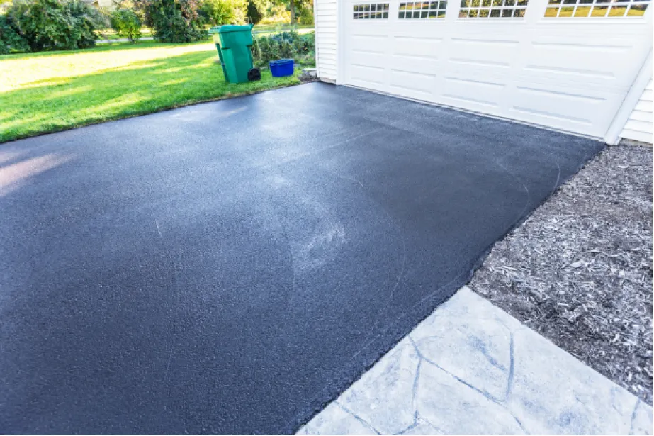 Freshly paved black asphalt driveway in front of a white garage, with green grass and trash bins nearby.