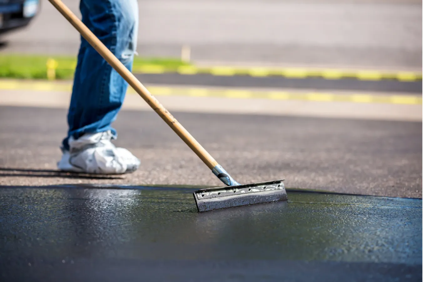 parking-sealcoating-1 Person spreading black sealant on asphalt with a squeegee tool; only legs and tool are visible.