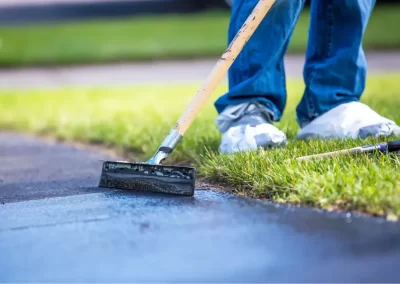 A Jones Asphalt VA expert applies black sealant to a driveway with a squeegee, wearing jeans and shoe covers, showcasing quality asphalt services.