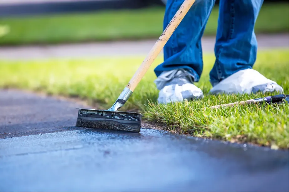 Person applying black sealant to driveway with a squeegee, wearing jeans and shoe covers, near green grass.