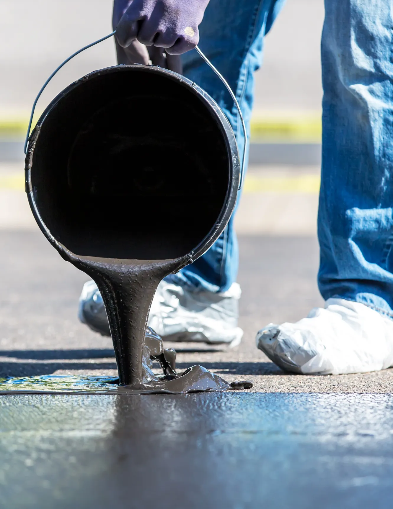 sealcoating-2 Person pouring black tar from a bucket onto pavement, wearing gloves and shoe covers, outdoors.