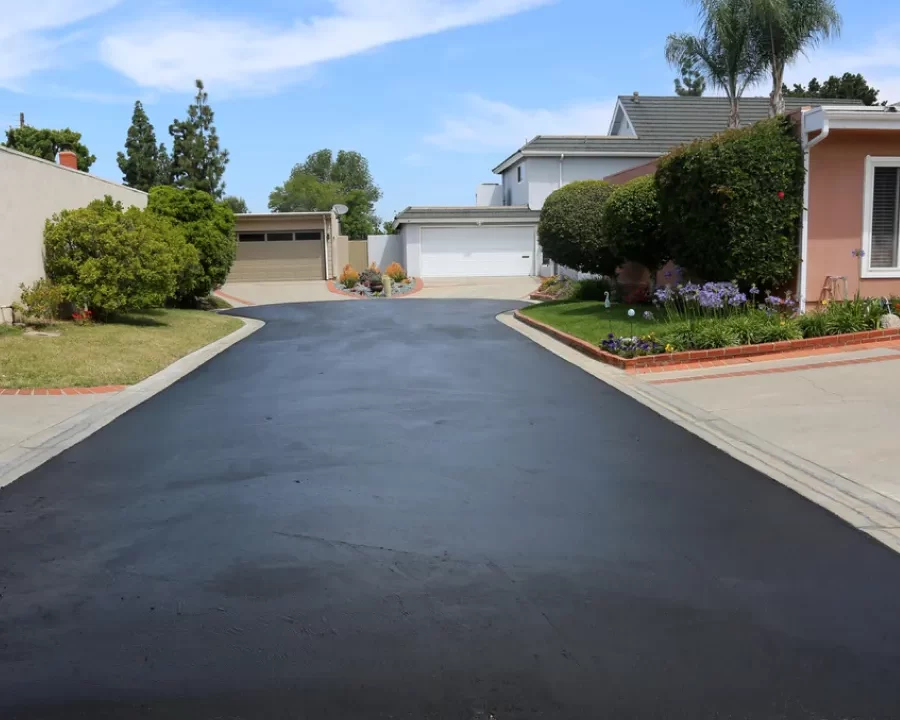 driveway-extensions-4-1 Freshly paved asphalt driveway in a suburban neighborhood with two houses, greenery, and blue sky.